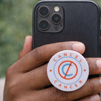 Hand holding a phone with a 'Cancel Cancer' phone holder on a blurred green natural background